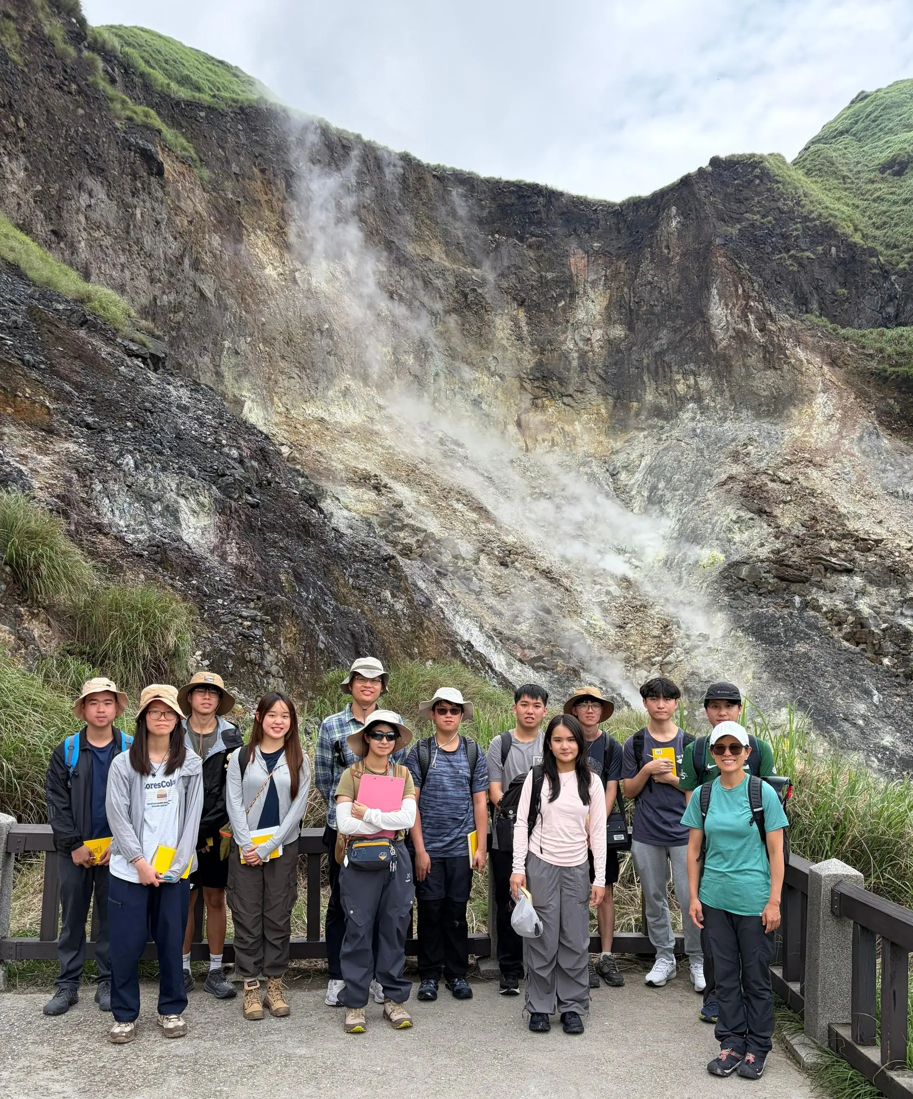 A group of students in sun hats standing on a walkway below a steaming, sulphur-stained hillside of active fumaroles.