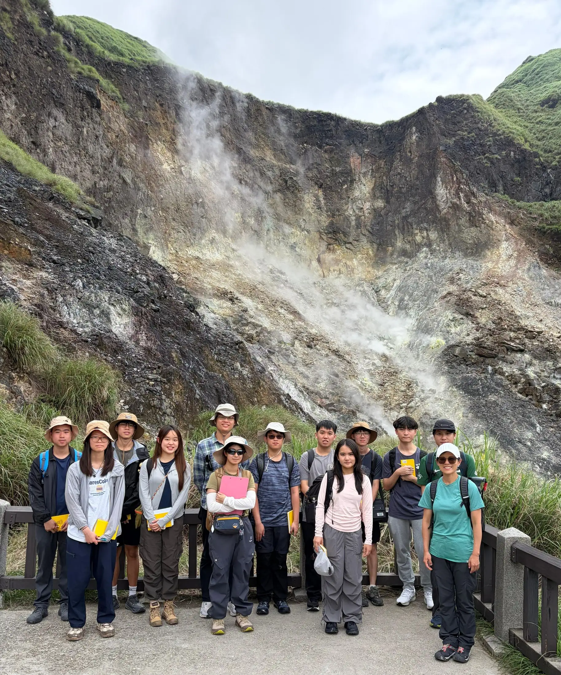 A group of students in sun hats standing on a walkway below a steaming, sulphur-stained hillside of active fumaroles.