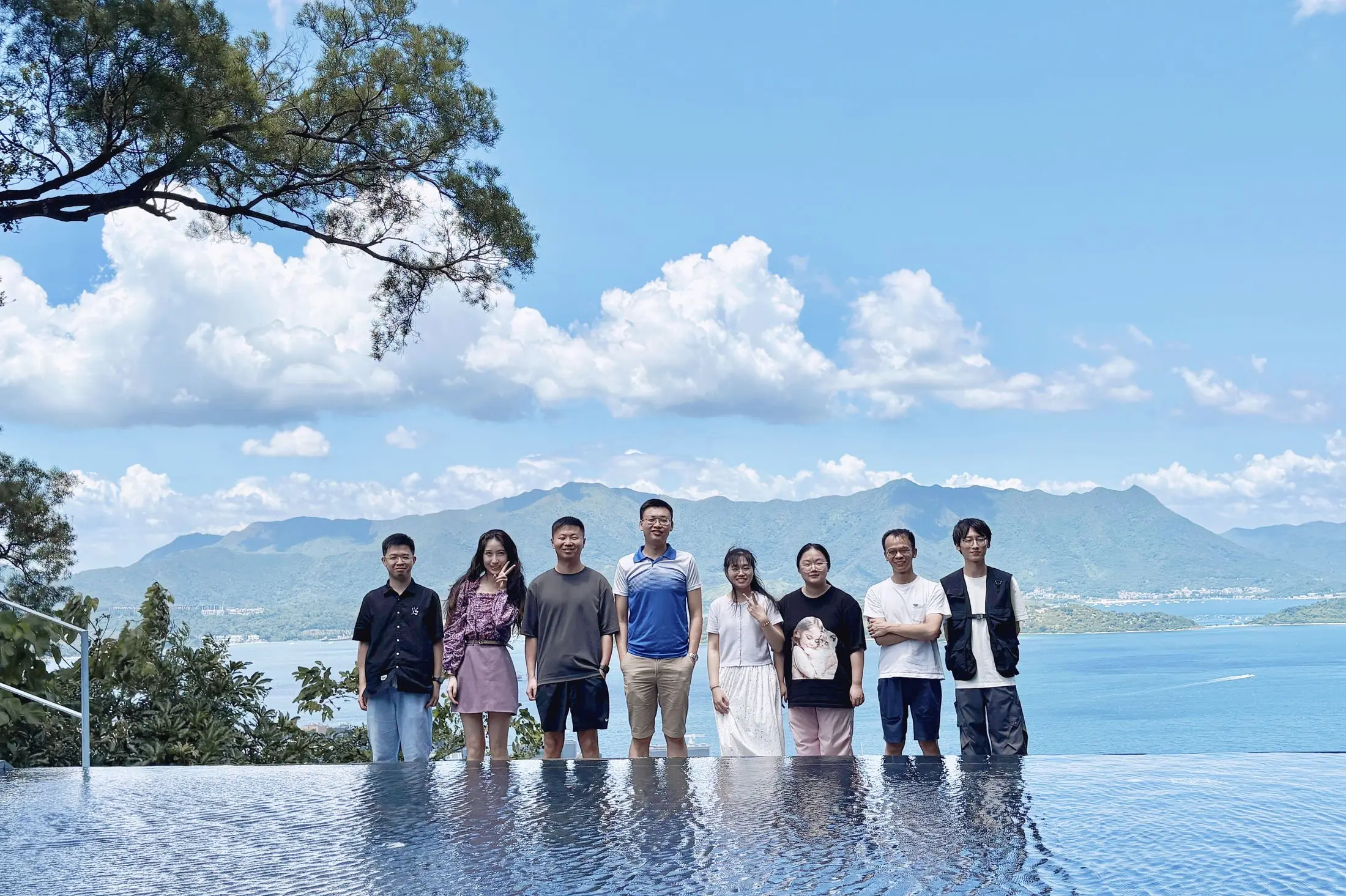 Ten group members standing in a shallow reflecting pool, with a volcanic lake and mountain ridge behind them.