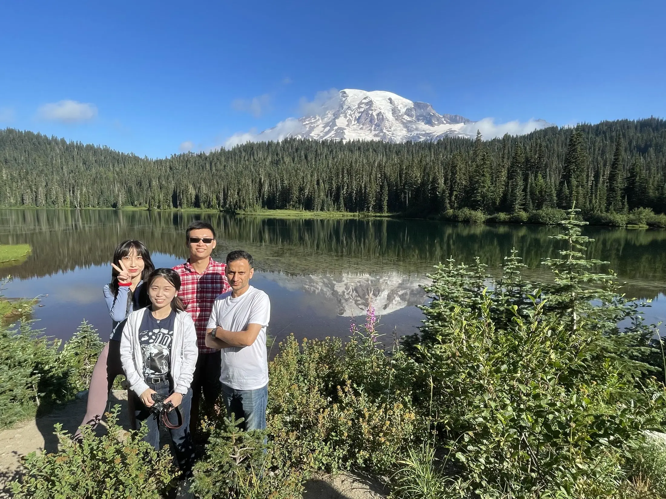 Three researchers beside a still lake that reflects a snow-capped stratovolcano framed by conifers.