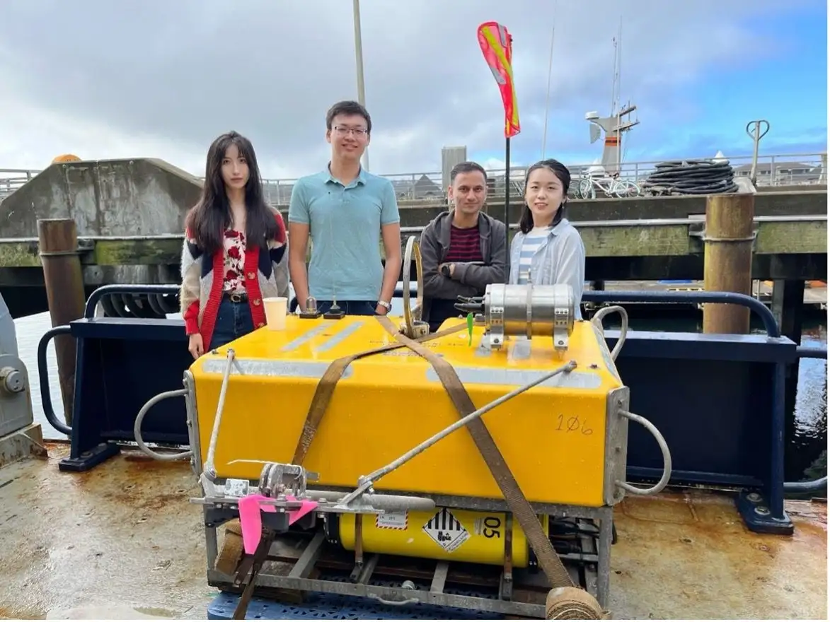 Four researchers behind a bright yellow ocean-bottom seismometer strapped to the deck of a research vessel.
