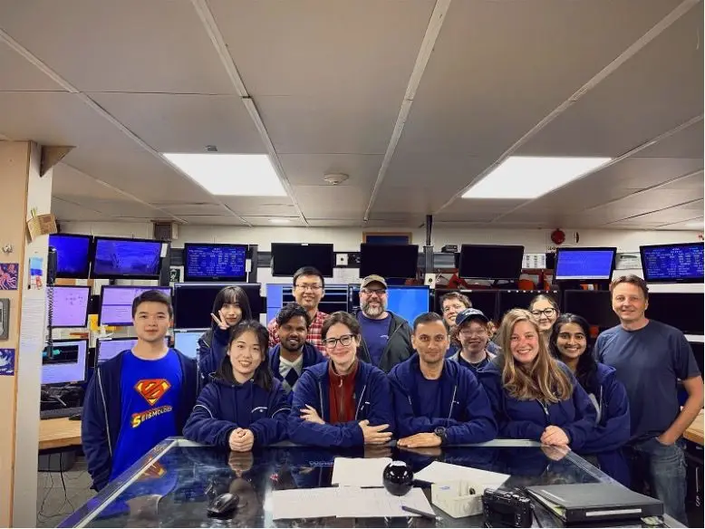 A science party of about a dozen people seated at a bank of monitors in a research vessel control room.