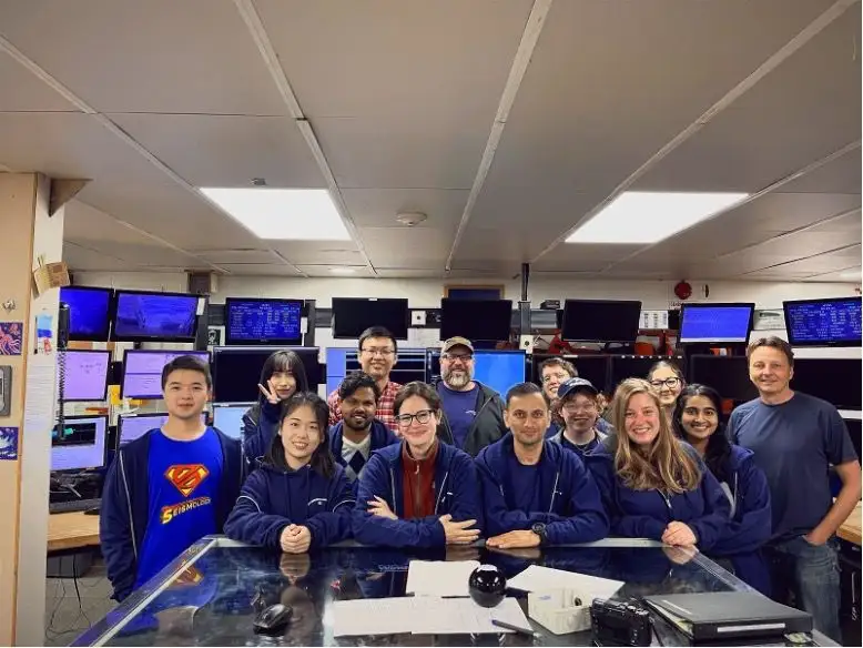 A science party of about a dozen people seated at a bank of monitors in a research vessel control room.