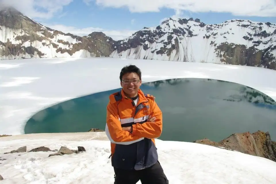 A researcher in orange field clothing standing at the rim of a turquoise crater lake ringed by snow.