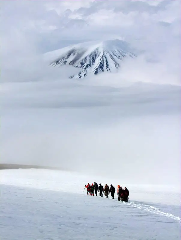 A line of researchers walking single file across a snowfield in dense fog beneath a dark volcanic cone.