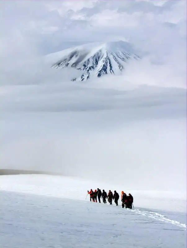 A line of researchers walking single file across a snowfield in dense fog beneath a dark volcanic cone.