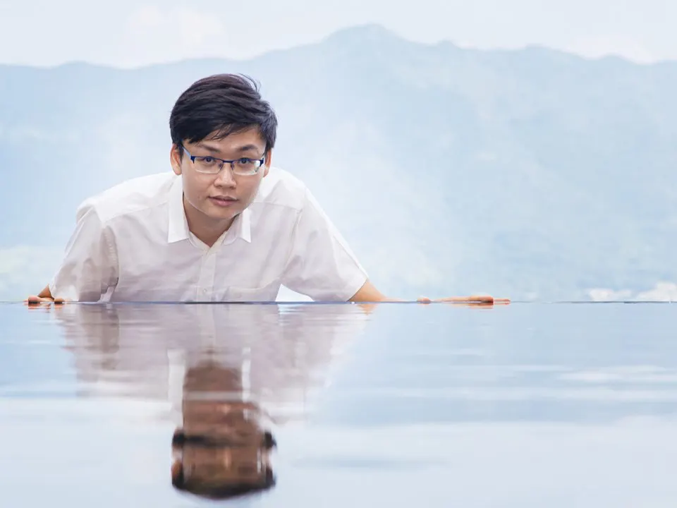 Professor Yen Joe Tan leaning over a mirror-still water surface that reflects him, with hazy mountains behind.