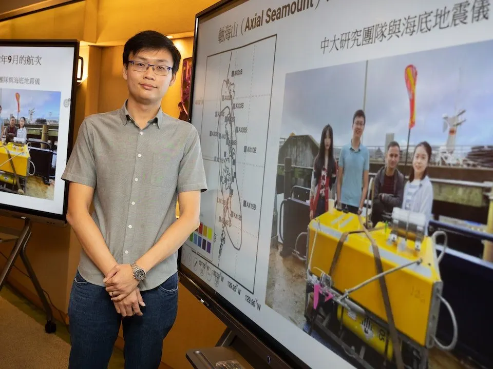 Professor Yen Joe Tan at a press briefing, standing before screens showing a yellow ocean-bottom seismometer and a map of Axial Seamount.