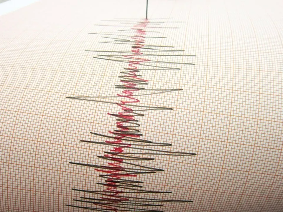 A seismograph needle scribbling a large earthquake in red and black onto ruled paper.